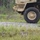 A tan/sand-colored military vehicle, likely an MRAP or similar tactical wheeled vehicle, is moving along a gravel road at speed, evidenced by motion blur on the large tire. A person in camouflage gear is visible lying prone in the vegetation on the opposite side of the road, partially concealed. The setting appears to be a military training area with a gravel road bordered by grass and sparse woodland.