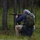 A person is kneeling in a forested outdoor setting while aiming a rifle. The individual is wearing a blue top, tan trousers, a black-and-white keffiyeh headscarf, and carrying an olive-green tactical backpack. The setting appears to be a sparse woodland with tall thin trees and ground-level vegetation.
