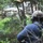 A person identified as 'HARPER' by a name tape on their cap is observed from behind, positioned among dense vegetation and trees. They are wearing tactical/protective ear muffs, a backpack with tactical straps, and a blue shirt. The subject appears to be observing a structure visible through the tree line.