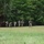 A group of approximately seven to eight individuals in military-style camouflage uniforms and protective face masks are moving across an open grassy field. They are carrying what appear to be training or simunition weapons, suggesting a military training exercise or tactical drill. A dense tree line forms the background behind the personnel.
