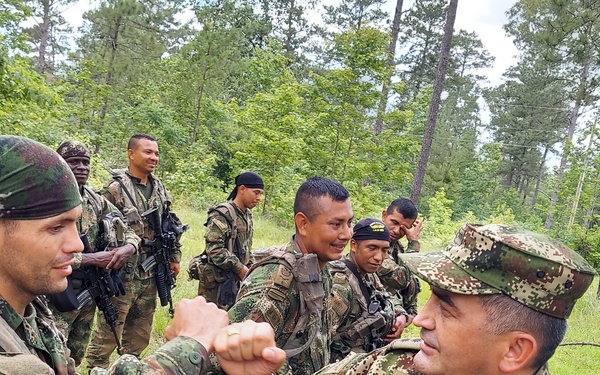 A group of military personnel in camouflage uniforms are gathered in a forested outdoor setting. Two soldiers in the foreground appear to be engaged in a fist-bump or handshake gesture, suggesting a positive interaction or greeting. Visible insignia and the word 'EJERCITO' (Spanish for 'Army') indicate these are Colombian Army soldiers.