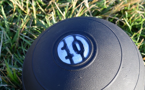 A black slam ball (medicine ball) resting on green grass outdoors. The ball displays a blue circular label with the number '10' visible on top, indicating its weight. The ball has a textured rubber surface with horizontal ridged lines around its circumference.