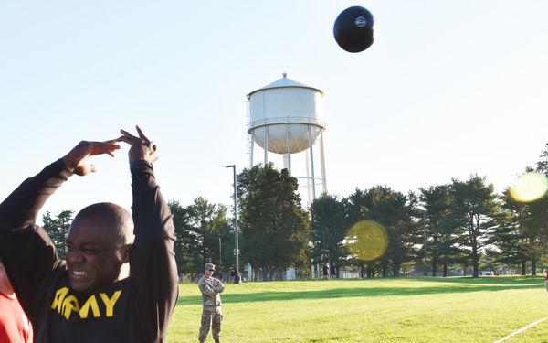 A soldier wearing an 'ARMY' branded black long-sleeve shirt is performing a standing medicine ball throw on a grassy field, likely as part of a physical fitness test. Several other military personnel in uniform and PT gear are visible observing or participating in the background. A white water tower and rows of trees are visible in the background of what appears to be a military installation.