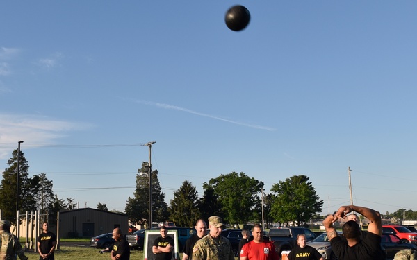 A group of U.S. Army soldiers and personnel are conducting a physical fitness assessment on a grass field at what appears to be a military installation. One soldier in the foreground is performing a standing power throw with a medicine ball, which is visibly airborne at the top of the frame. Observers, scorers with clipboards, and a whiteboard scoring station are present in the background.