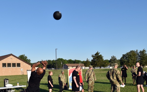 Military personnel are conducting a physical fitness assessment on a grass field, with one soldier performing a standing power throw using a medicine ball. The event appears to be part of the Army Combat Fitness Test (ACFT), with observers in both PT (physical training) uniforms and Army Combat Uniforms (ACU) present. Orange cones and white rope mark the testing lane, and a unit guidon flag is visible in the background.