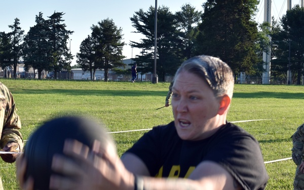 A military fitness test is being conducted on a grass field, with a female soldier in Army PT (physical training) black shirt throwing or pushing a medicine ball. A uniformed soldier with a clipboard observes and records the performance, while additional personnel are visible in the background. The setting appears to be a military installation based on the uniforms and organized testing setup.