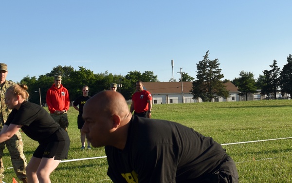 Military personnel are participating in a physical fitness assessment or training exercise on a grass field. Two soldiers in black PT (physical training) uniforms are performing what appears to be a medicine ball throw event, while observers in both military camouflage uniforms and red athletic shirts watch. The setting appears to be a military installation based on the uniforms, flags, and background buildings.