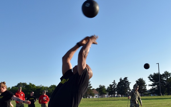 Military personnel are conducting a physical fitness test on a grass field, performing a standing power throw with medicine balls. The event appears to be part of the Army Combat Fitness Test (ACFT), with scorers and observers present. The setting is a military installation identifiable by the water tower and uniformed personnel.