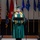 Idaho Youth Challenge Graduates 15th Class A graduate in a green cap and gown with a gold honor stole stands at a podium delivering a speech during a graduation ceremony. Multiple flags are displayed in the background, including the United States flag and what appear to be military branch flags. The setting appears to be an indoor auditorium or ceremonial hall.