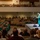 Idaho Youth Challenge Graduates 15th Class A graduation ceremony is taking place in a large auditorium or event hall. A student in green cap and gown with a yellow stole stands at a podium on a lit stage, addressing an audience that includes rows of fellow graduates in matching green regalia and seated family members. The venue features both main floor and balcony seating, with a projection screen visible on the right side of the stage.