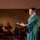 Idaho Youth Challenge Graduates 15th Class A graduate wearing a green cap and gown stands at a microphone stand delivering a speech, likely a valedictorian or commencement address. The speaker holds papers or a clipboard and gestures with one hand toward an audience seated in the background. The setting appears to be an auditorium or large hall during a graduation ceremony.
