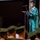 Idaho Youth Challenge Graduates 15th Class A young male graduate in a green cap and gown stands at a podium with a microphone, delivering a speech during a graduation ceremony. He appears to be a student speaker, likely a valedictorian or class representative, addressing fellow graduates seated in rows in the foreground. The setting is an indoor auditorium or event hall.