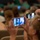 Idaho Youth Challenge Graduates 15th Class An audience seated in a venue is capturing a live event or performance using smartphones. The foreground shows a woman with blonde hair and glasses holding up a teal-colored smartphone, with its screen displaying what appears to be a stage performance. Multiple other audience members are also visible holding up phones to record or photograph the event.
