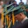 Idaho Youth Challenge Graduates 15th Class A row of graduates wearing green mortarboard caps and gowns with gold/yellow tassels are bowing their heads, likely during a prayer or moment of reflection at a graduation ceremony. The image is taken from a side angle, showing multiple graduates in a line receding into the background. The foreground graduate is in sharp focus while the background graduates become progressively blurred.