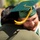 Idaho Youth Challenge Graduates 15th Class A graduate wearing a green cap and gown with a gold tassel and gold stole is sharing a warm embrace with another person at what appears to be an outdoor graduation ceremony. The graduate is smiling broadly and wearing braces, visibly expressing joy and celebration. Other graduates in green caps and gowns are visible in the blurred background.