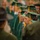 Idaho Youth Challenge Graduates 15th Class A graduation ceremony is underway with a large group of graduates wearing green caps and gowns with gold/yellow tassels. The crowd of graduates appears to be standing in rows, likely during a processional or ceremony. The image is shot with a shallow depth of field, with one graduate's face in sharp focus amid a blurred crowd.