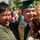 Idaho Youth Challenge Graduates 15th Class A graduation ceremony is taking place outdoors, with a young graduate in a green cap and gown being embraced by two people, likely family members. The graduate is smiling broadly and holding a green diploma cover while a man to his left laughs joyfully. The background shows other graduates and attendees in a sunny outdoor setting.