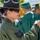 Idaho Youth Challenge Graduates 15th Class A military service member wearing an olive green flight suit with a Technical Sergeant rank insignia patch and a campaign/drill sergeant hat is present at what appears to be a graduation ceremony. The service member is interacting with graduates dressed in green caps and gowns with gold tassels. The scene suggests a celebratory moment, possibly a fist bump or handshake between the military member and a graduate.
