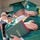 Idaho Youth Challenge Graduates 15th Class Two graduates in green caps and gowns share an emotional embrace at what appears to be a graduation ceremony. One graduate is visibly holding a dark red diploma cover with 'of Graduation' text visible. The scene captures a candid, heartfelt moment surrounded by other graduates and attendees in the background.
