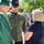 Idaho Youth Challenge Graduates 15th Class An outdoor graduation ceremony scene featuring a graduate in green cap and gown conversing with an Idaho Department of Fish and Game (IDYCA) officer named Brown and a woman in a navy blue shirt. The setting appears to be a sunny day with other graduates and attendees visible in the background. The interaction appears casual and friendly, likely a post-ceremony gathering.