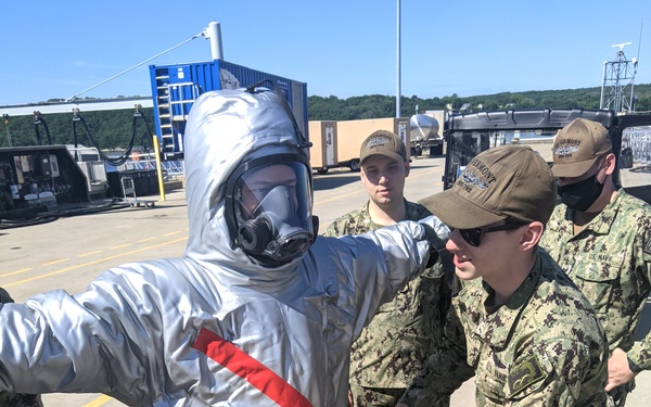 U.S. Navy personnel are conducting a HAZMAT or CBRN (Chemical, Biological, Radiological, Nuclear) training exercise at what appears to be a naval installation. One service member is wearing a silver aluminized protective suit with a full-face respirator/gas mask and yellow boot covers, while others in Navy Working Uniform (NWU Type III) assist with fitting or inspecting the suit. Several sailors wearing caps with what appears to be 'USS Vermont' insignia are observing the procedure.