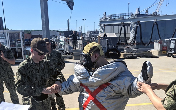 Military personnel in Navy Working Uniforms (NWU) are assisting a colleague in donning a proximity/fire proximity suit with a gas mask at what appears to be a naval installation pier. One sailor holds a checklist document while others help secure the protective suit on the individual. The setting is an outdoor pier area with large naval vessels visible in the background.