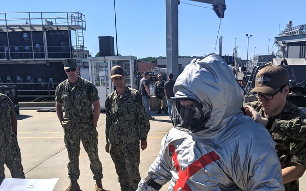 Military personnel in camouflage uniforms are gathered outdoors at what appears to be a naval facility or port. One individual is wearing a silver hazmat/CBRN protective suit with a red X marking on the back, while others assist with donning the equipment. The scene appears to be a training exercise or demonstration involving chemical, biological, radiological, or nuclear (CBRN) protective gear.
