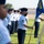 A formal military parade or ceremony featuring U.S. Air Force personnel in service dress uniforms standing in formation on a grass field. A central figure wearing a black face mask holds a blue guidon flag bearing the text '37 ABW' (37th Air Base Wing). The scene suggests a unit formation or change of command ceremony conducted during the COVID-19 pandemic era.