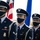 A U.S. Air Force Base Honor Guard stands at attention during a formal ceremony, wearing dress uniforms and COVID-19 face masks. The color guard is carrying the American flag and what appears to be an Air Force unit flag. The personnel display various rank insignia and service ribbons on their uniforms.