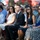 A group of civilians and what appears to be military family members are seated outdoors attending a formal ceremony. The audience is dressed in business and semi-formal attire, with some individuals holding printed programs. The setting appears to be an outdoor military or official ceremony based on uniformed personnel visible in the background.
