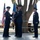 A formal U.S. Air Force change of command ceremony is taking place on an outdoor stage. Two senior officers are exchanging a unit guidon (flag), which is the traditional symbol of command transfer, while a third officer stands at attention to the right. The ceremony appears to be conducted at an air base, with aircraft and military equipment visible in the background.
