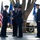 A formal U.S. Air Force change of command ceremony is taking place on an outdoor stage. Two senior officers are exchanging a unit guidon (flag), which is the central symbolic act of a change of command. Additional uniformed personnel stand at attention nearby, with U.S. and Air Force flags displayed prominently.