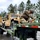 1041st Transportation Company regains identity during eventful annual training A U.S. Army soldier in OCP (Operational Camouflage Pattern) uniform is securing a HMMWV (Humvee) onto a military flatbed trailer using tie-down straps. The vehicle, marked '1041 TC', is loaded on a green military low-boy trailer in what appears to be a wet, overcast outdoor environment. A second soldier is visible in the background near additional military vehicles.