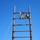 1041st Transportation Company regains identity during eventful annual training Two individuals in military/camouflage uniforms are seated at the top of a tall wooden ladder structure against a clear blue sky. One person is holding up a red and yellow flag, suggesting a completion or achievement moment. The structure appears to be a military obstacle course or training apparatus stabilized by guy-wires.