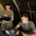 Two military personnel in olive drab t-shirts are working inside what appears to be a military field tent or temporary command post. One individual wearing camouflage pants and glasses is standing and holding a tablet device, while the other is seated at a table operating a laptop computer. The setting suggests a field operations or command and control environment.