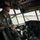 Connecticut Guard upgrades C-130H fleet A military aviator in an olive drab flight suit sits in the cockpit of a large military transport aircraft, appearing to be conducting pre-flight checks or adjusting controls. The cockpit features an extensive array of analog and digital instruments, switches, and overhead panels typical of a large cargo aircraft such as a C-130 Hercules. The aircraft is positioned on a tarmac with a runway and tree line visible through the windscreen.