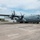 Connecticut Guard upgrades C-130H fleet A U.S. Air Force C-130 Hercules transport aircraft with tail number 7322 is parked on a flight line apron at a military airbase. Ground personnel in military uniforms are conducting pre-flight or maintenance activities around the aircraft. The setting appears to be a warm, partly cloudy day with base infrastructure and trees visible in the background.