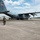 Connecticut Guard upgrades C-130H fleet A U.S. Air National Guard C-130 Hercules transport aircraft (tail number 67322) is parked on a tarmac. A flight crew member in an olive green flight suit walks away from the aircraft carrying equipment, while additional personnel are visible near the rear cargo ramp. The setting appears to be a military airfield with hangars visible in the background.