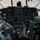 Connecticut Guard upgrades C-130H fleet The image shows the interior cockpit of a large military transport aircraft, likely a C-130 Hercules or similar aircraft, viewed from behind the two pilot seats. The cockpit features an extensive analog instrument panel with numerous gauges, dials, switches, and controls typical of a mid-to-late 20th century military aircraft. The aircraft appears to be parked on a tarmac, visible through the large forward-facing windshield panels.