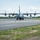 Connecticut Guard upgrades C-130H fleet A military turboprop transport aircraft, consistent with a C-130 Hercules, is taxiing on an airport taxiway or runway facing the camera head-on. All four propellers are spinning, visible as motion-blurred discs, indicating the engines are running at operational power. The setting appears to be a military or joint-use airfield with standard taxiway markings and orange/blue marker cones.