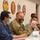 A group of individuals, including at least one U.S. Army soldier in OCP (Operational Camouflage Pattern) uniform, are seated at a conference table during what appears to be an official military briefing or meeting. All participants are wearing face masks, suggesting COVID-19 era protocols. The setting is a formal military conference room decorated with unit insignia patches and a U.S. flag.