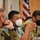 Three individuals are seated at a table during what appears to be a formal military meeting or briefing. Two are wearing military camouflage uniforms, while one in the center wears civilian attire. All three are wearing face masks, consistent with COVID-19 era protocols.