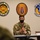A U.S. Army Colonel wearing a black face mask and OCP camouflage uniform is seated at a table, apparently speaking or briefing an audience. A name placard in front of him reads 'COL Christopher O'Gwin.' Three military unit insignia plaques are mounted on the wall behind him, including one for Soto Cano Air Base Honduras and the 612th Air Base Squadron.