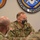 A U.S. Army Colonel (identified by eagle insignia on collar) with the name tape 'GVENTER' is seated at a meeting or briefing, wearing OCP (Operational Camouflage Pattern) uniform and a camouflage face mask. He is engaged in conversation with other military personnel. The setting appears to be an official military facility, with organizational seals/emblems visible on the wall behind him.