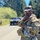 A U.S. Army soldier in full Operational Camouflage Pattern (OCP) uniform stands on a gravel road during what appears to be a field training exercise. The soldier is wearing tactical gear including a plate carrier, patrol cap, and reflective eyewear, and appears to be adjusting equipment or holding a radio. Military vehicles, including at least one HMMWV (Humvee), are visible in the background along a tree-lined road.