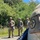 The image shows four military personnel in OCP (Operational Camouflage Pattern) uniforms during what appears to be a field training exercise. Three soldiers stand on a gravel path in front of dense green vegetation, while a fourth soldier in the foreground is partially visible with a rifle barrel extending into frame. A unit patch is visible on the foreground soldier's shoulder, appearing to be a hexagonal insignia.