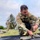 A U.S. Army soldier in Operational Camouflage Pattern (OCP) uniform is crouched down on a grassy field, working with what appears to be a tent or tarp being set up or broken down. He is wearing a plate carrier/tactical vest and has a U.S. flag patch and a unit patch visible on his left shoulder. At least one other soldier is visible in the background near military vehicles.