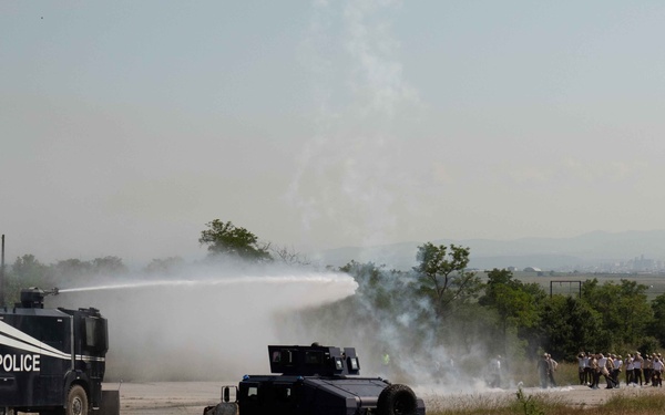 The image shows a law enforcement training exercise or demonstration involving riot control vehicles and personnel. A marked 'POLICE' water cannon vehicle is actively discharging a high-pressure water stream toward a group of people on the right side of the frame. A second armored tactical vehicle is also visible in the foreground, and smoke or gas is present in the scene.