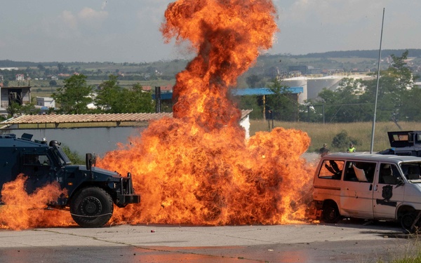 A large controlled explosion or pyrotechnic detonation is occurring between military/law enforcement armored vehicles and civilian vehicles on a paved surface. The scene appears to be a training exercise or demonstration, evidenced by the staged arrangement of vehicles and a visible safety observer in a yellow vest in the background. The explosion produces a tall column of orange flame rising several meters into the air.