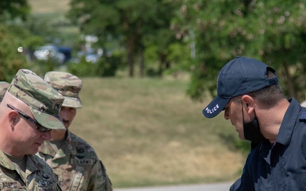 The image shows U.S. Army soldiers interacting with what appears to be a Kosovo Police officer during what looks like a joint training or liaison event. The police officer is handling equipment on a tripod, possibly a spotting scope, while a soldier identified by the name tape 'ADAMS' observes. Additional U.S. Army personnel are visible in the background.